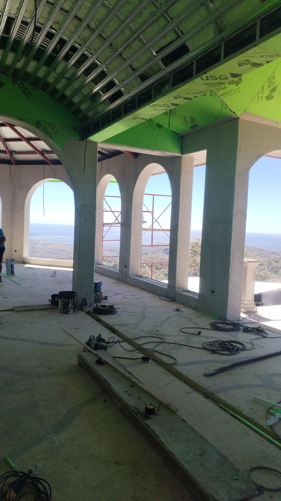 Una construcción en proceso con arcos que ofrecen vista al mar y paneles de yeso verde en el techo.