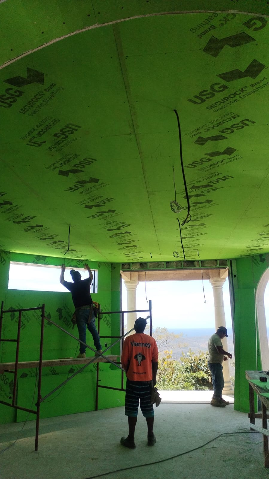 Trabajadores de construcción instalan paneles de yeso verdes en el techo y paredes de una estructura en obra.